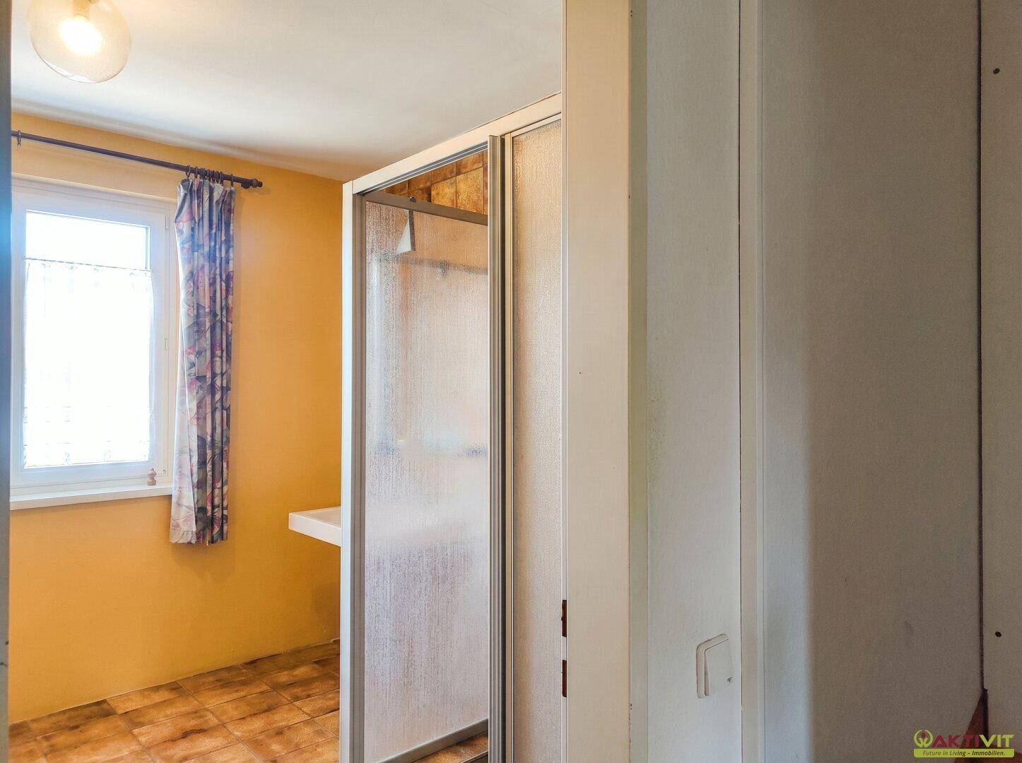 Gelb gestrichener Raum mit Fliesenboden und einer Duschkabine neben dem Fenster.