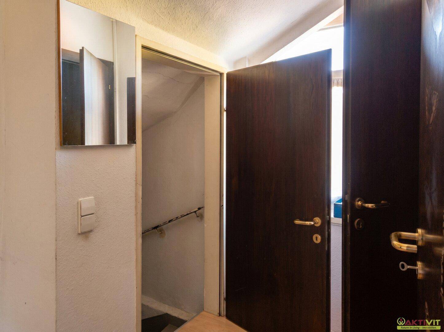 Eingangsbereich mit dunkler Holztür, einem Spiegel und Zugang zu einer Treppe.