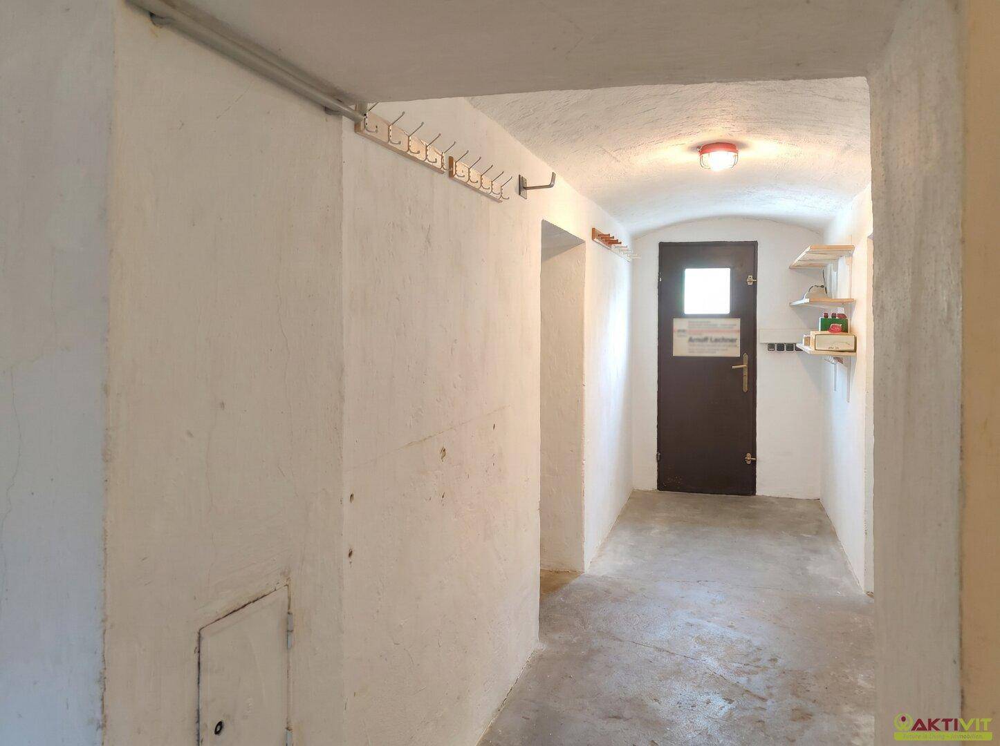 Langer Kellerflur mit rauen weißen Wänden, Betonboden und einer dunklen Tür am Ende.
