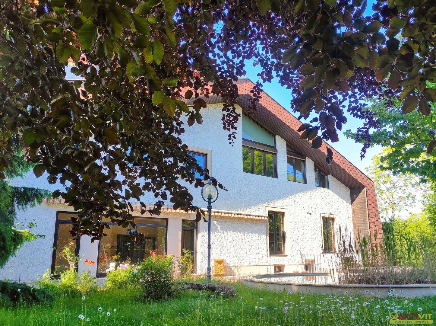 Attraktives Haus mit weißer Fassade und großen Fenstern, umgeben von einem grünen Garten mit Teich.