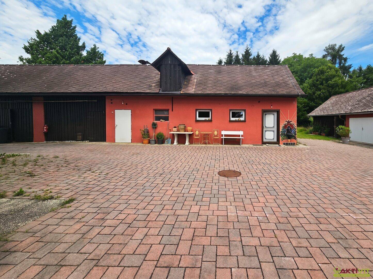 Rotes Gebäude mit mehreren Eingängen und gepflastertem Hof, umgeben von Bäumen und Natur.