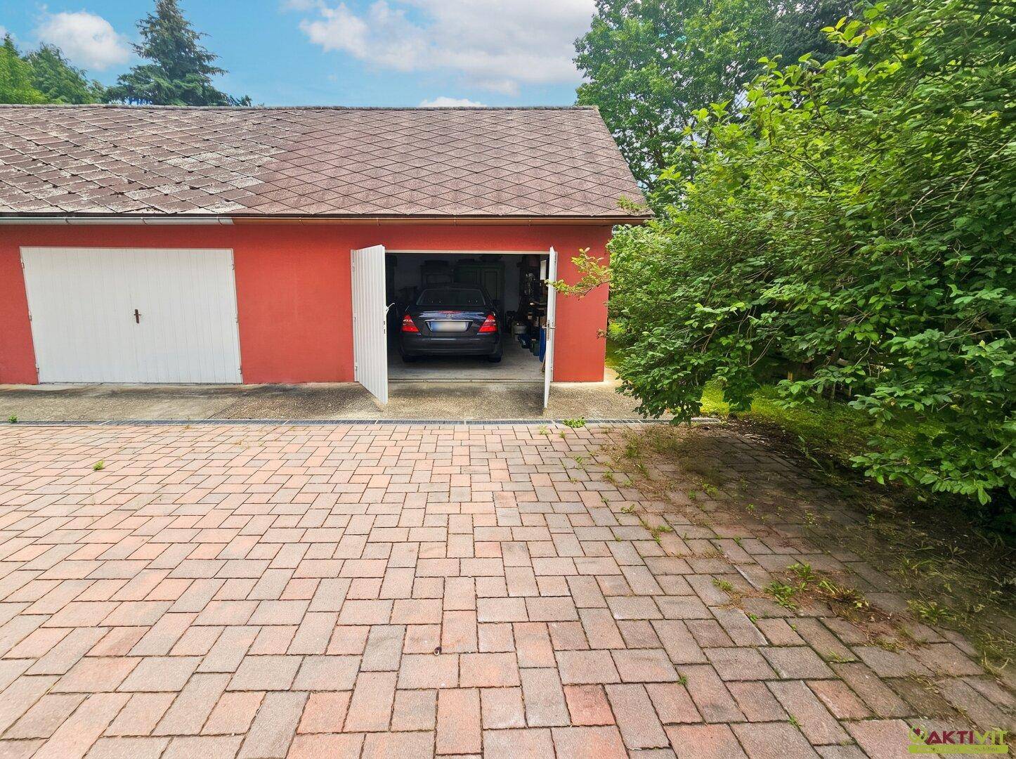 Rotes Garagengebäude mit offenem Tor und gepflastertem Vorplatz, umgeben von grünen Büschen.