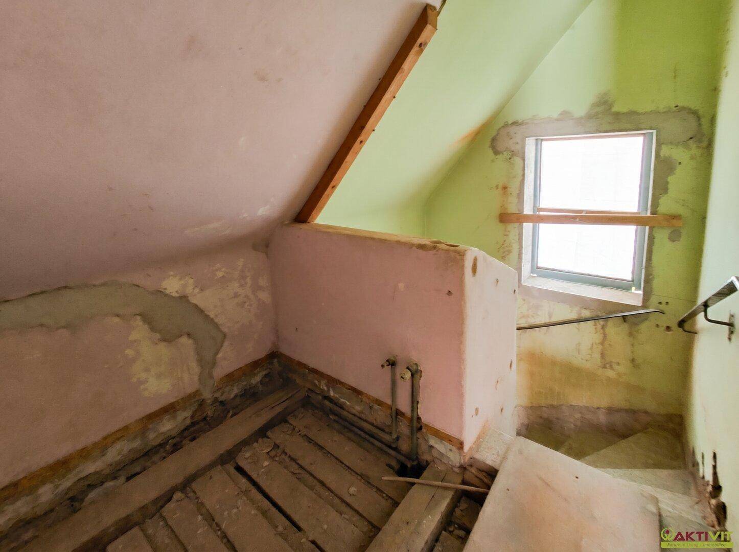 Unfertiger Dachbodenbereich mit schrägen Wänden, Fenster und sichtbaren Holzböden.