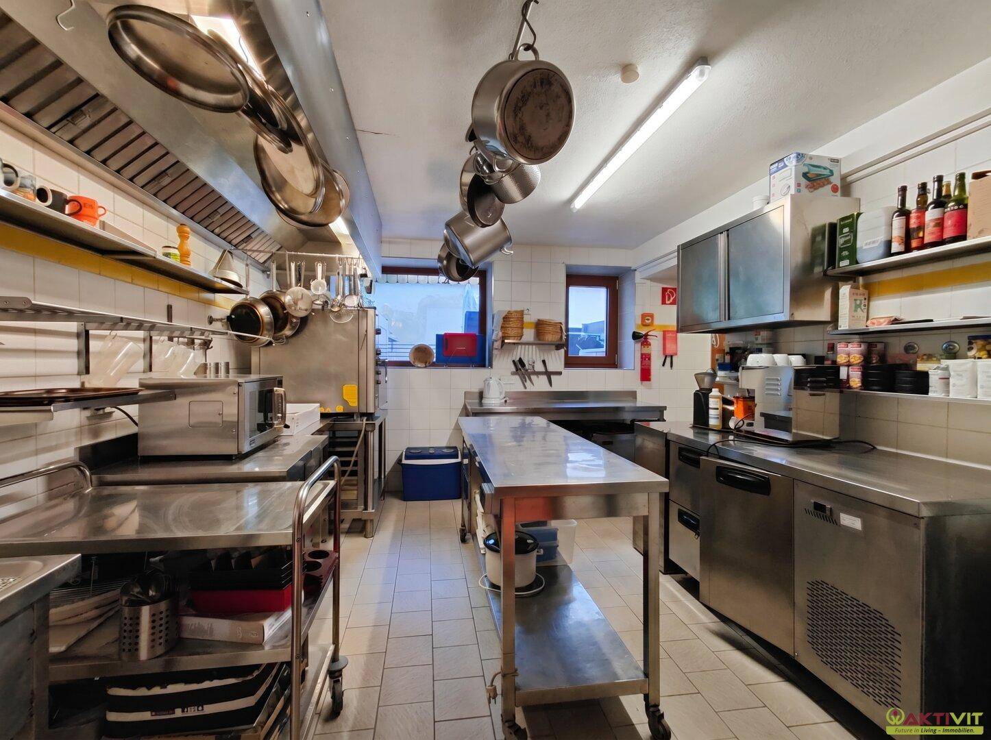 Große Gewerbeküche mit zentralem Arbeitstisch, vielen Edelstahlgeräten und Fliesenboden.