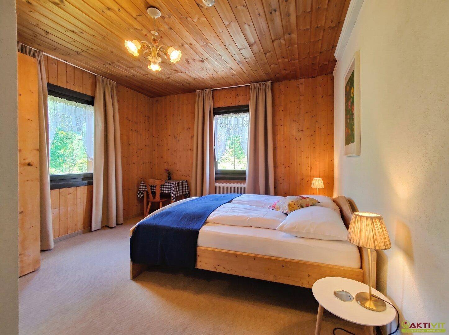 Gemütliches Schlafzimmer mit Holzwänden und -decke, Doppelbett und zwei Fenstern mit Blick ins Grüne.