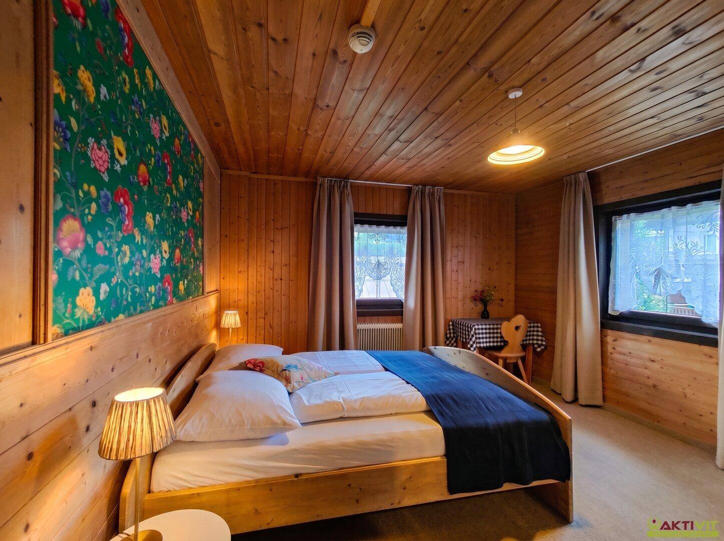Schlafzimmer mit Holzwänden, Decke und einem farbenfrohen Blumenmuster an der Wand hinter dem Doppelbett.