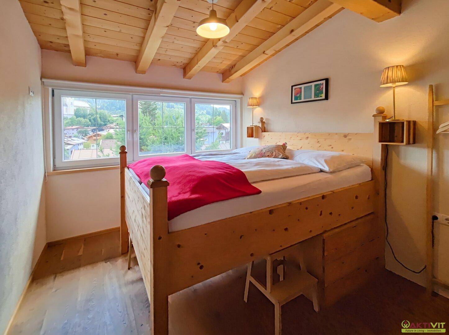 Helles Schlafzimmer mit erhöhtem Holzbett, Holzbalkendecke und großen Fenstern mit Blick ins Grüne.