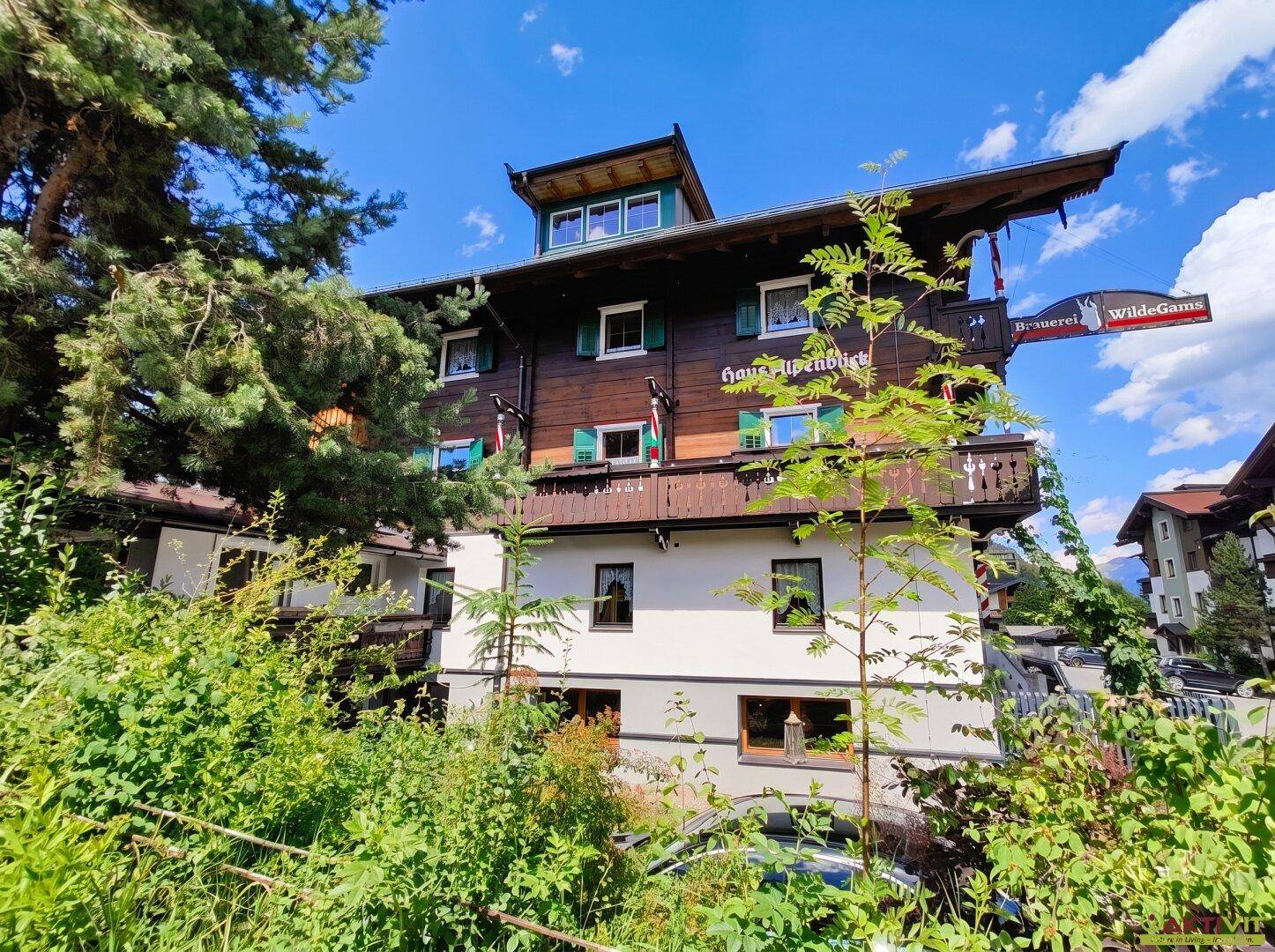 Traditionelles Gebäude im Alpenstil, umgeben von üppigem Grün unter blauem Himmel.