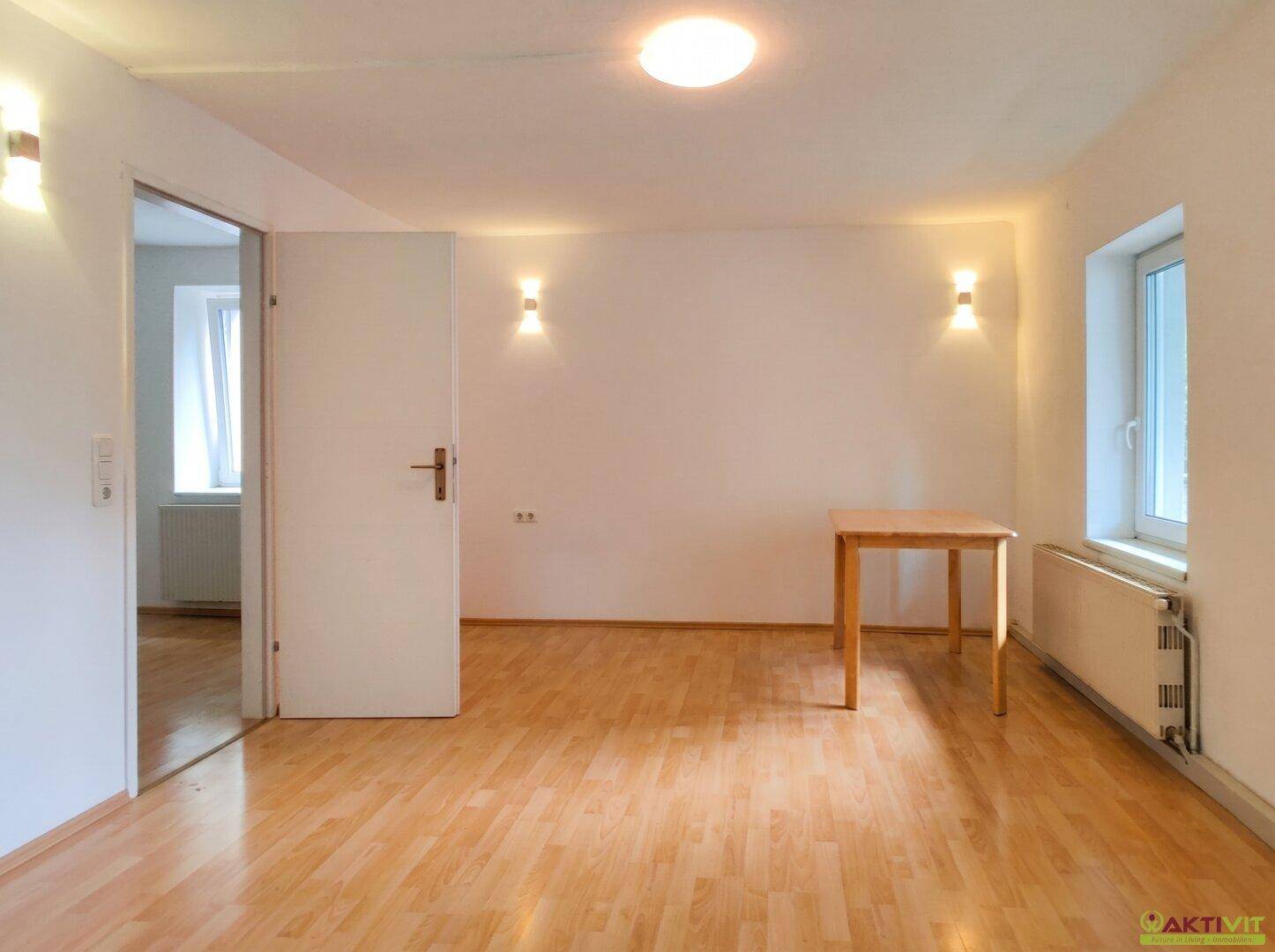 Helles Wohnzimmer mit Holzboden, weißen Wänden und einem kleinen Holztisch am Fenster.