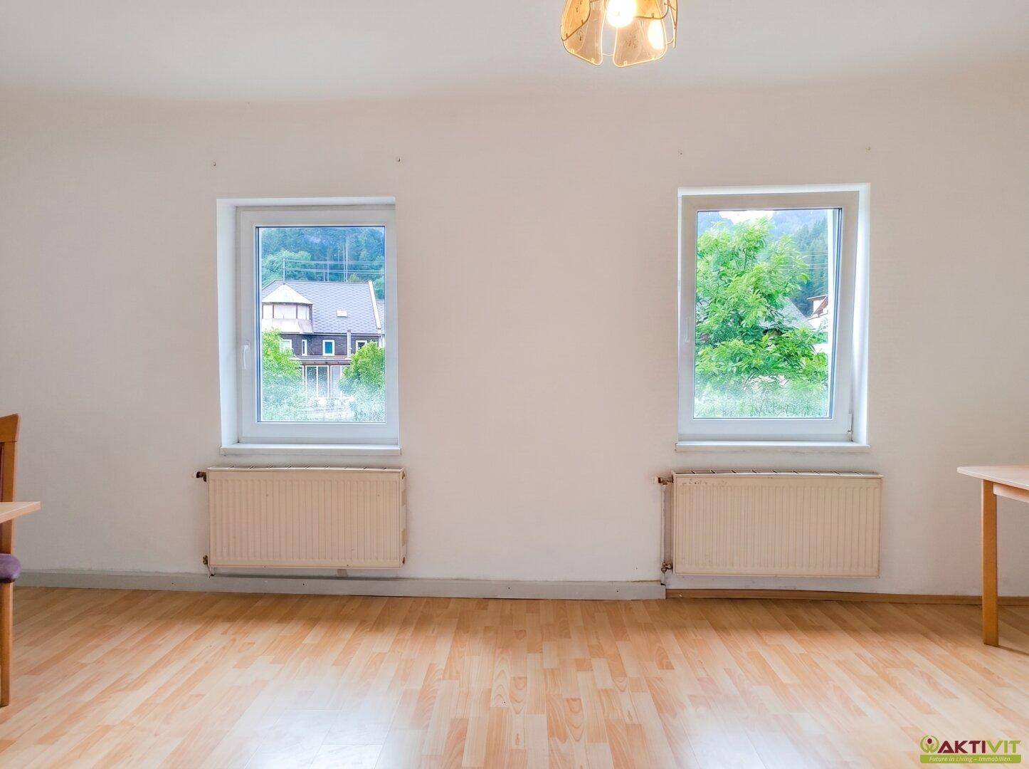 Wohnzimmer mit Holzboden und zwei Fenstern, die einen weiten Blick ins Grüne bieten.