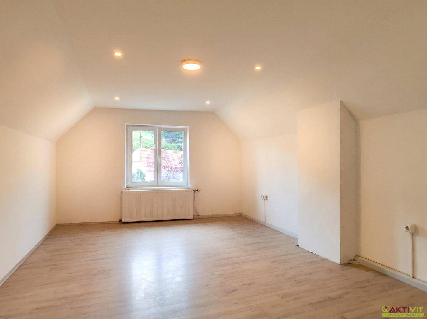 Geräumiger Dachbodenraum mit hellem Holzboden, schrägen Decken und einem Fenster.