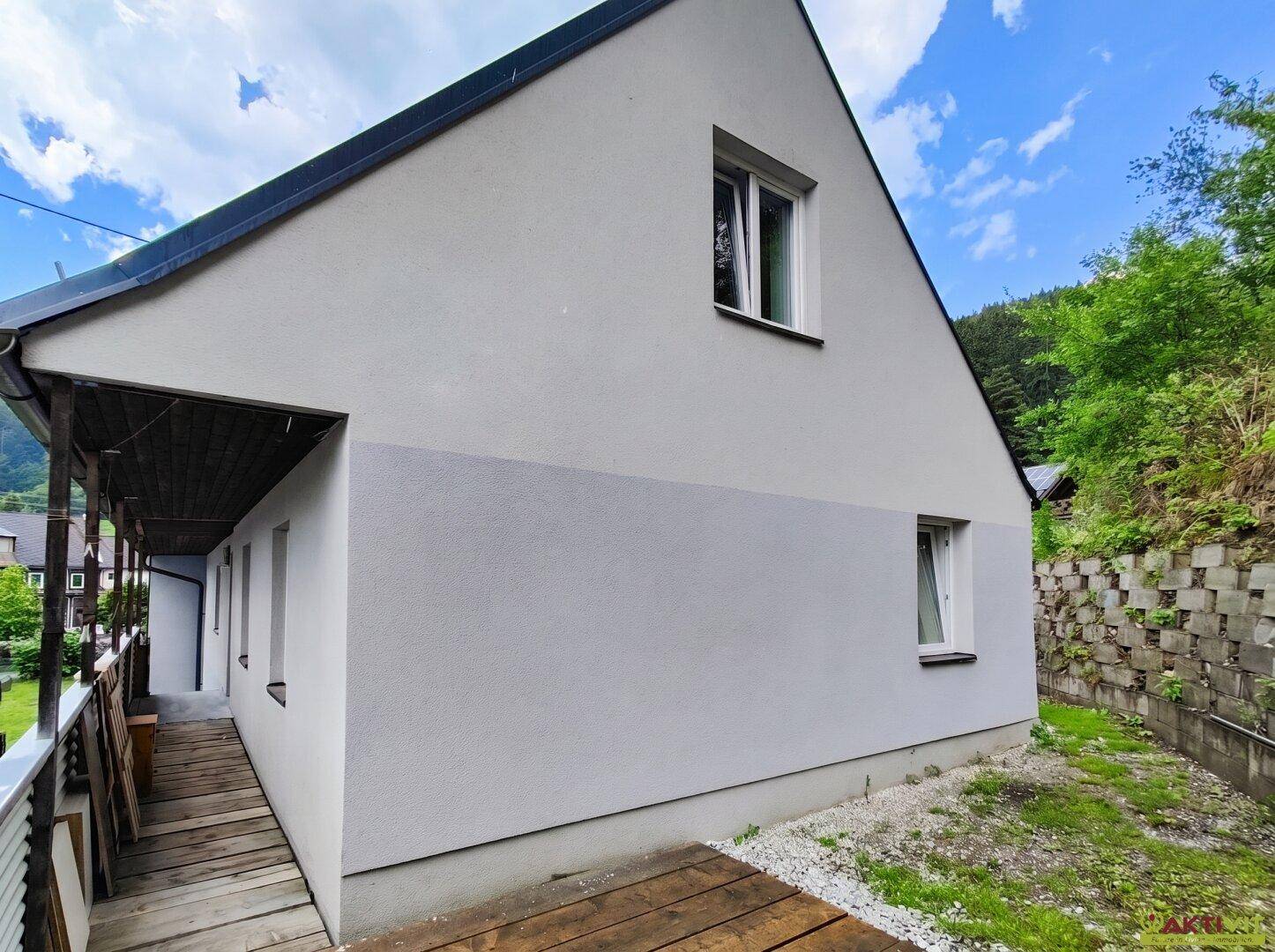 Seitenansicht des Hauses mit einer Holzveranda und einer Stützmauer im Hintergrund.