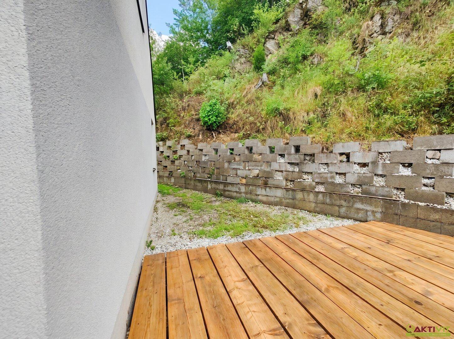 Gartenbereich mit einer Holzterrasse und einer gemauerten Stützmauer im Hintergrund.