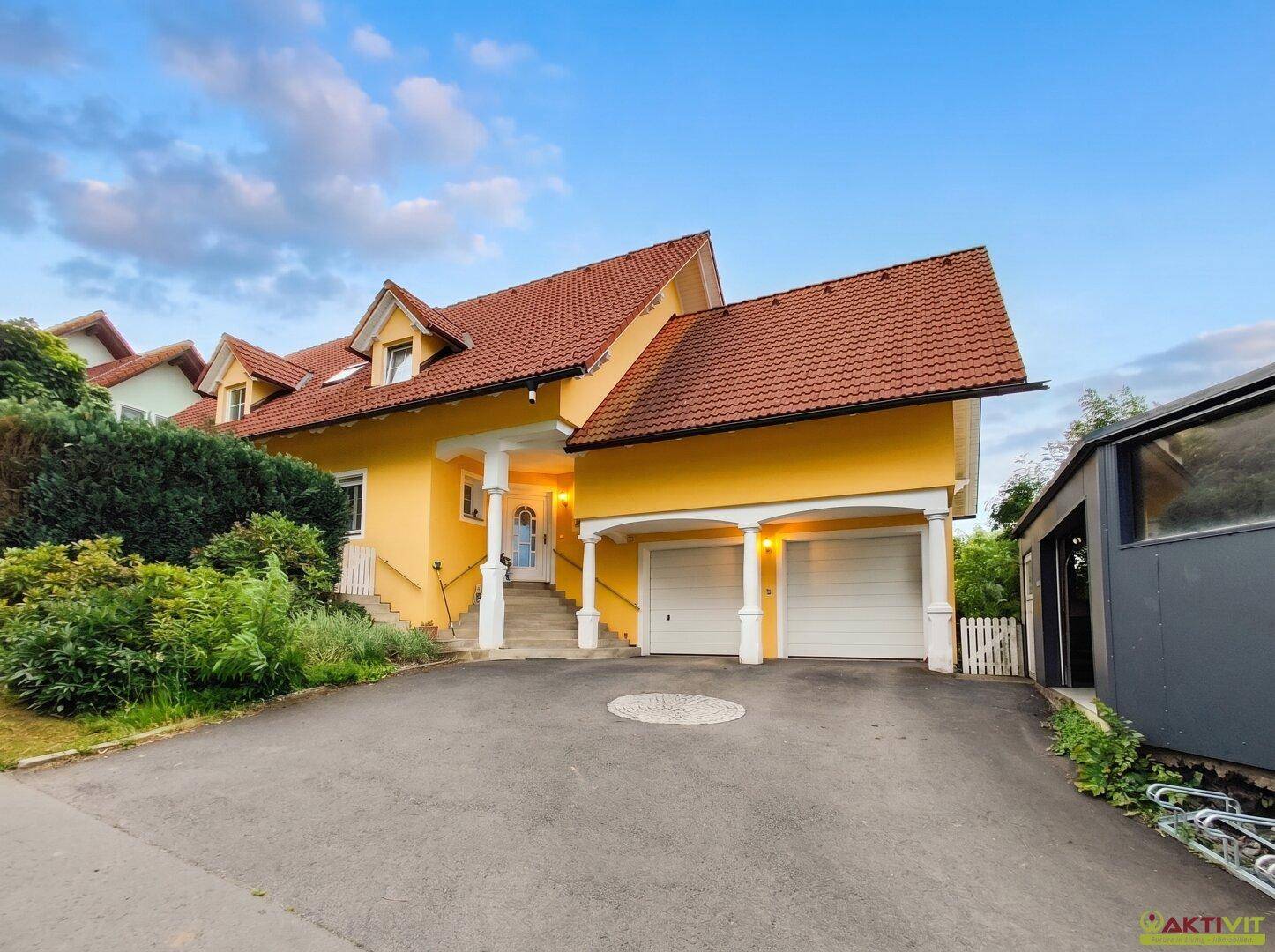 Vorderansicht des gelben Hauses mit Doppelgarage und gepflasterter Auffahrt unter Abendhimmel.