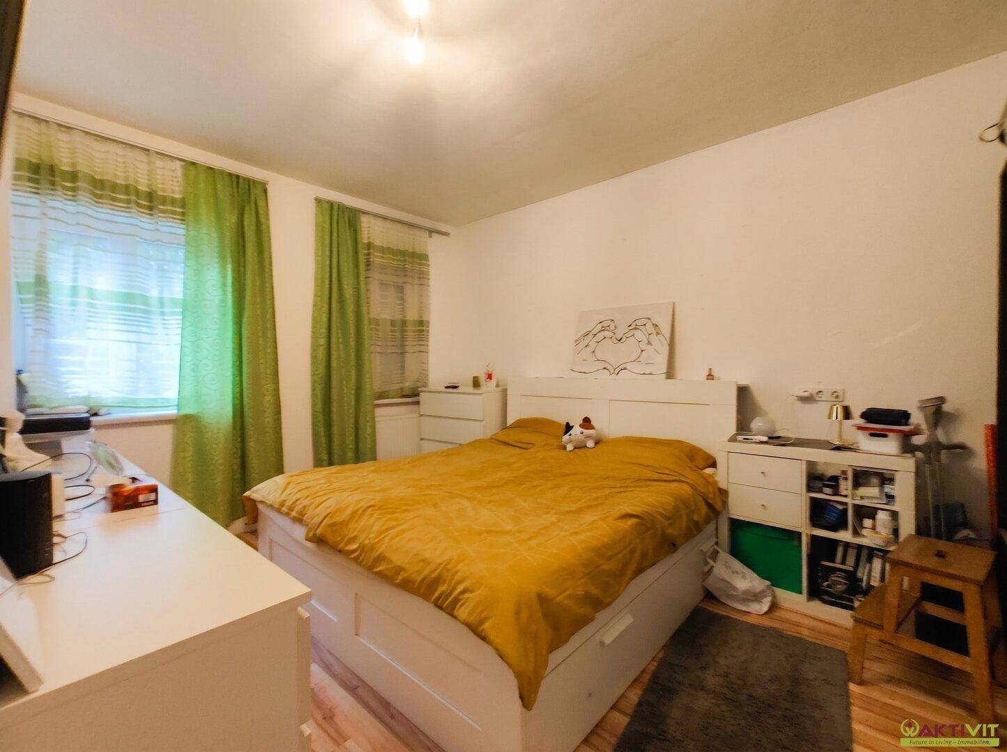 Schlafzimmer mit Doppelbett, weißen Möbeln und grünem Vorhang, hell durch Fenster.