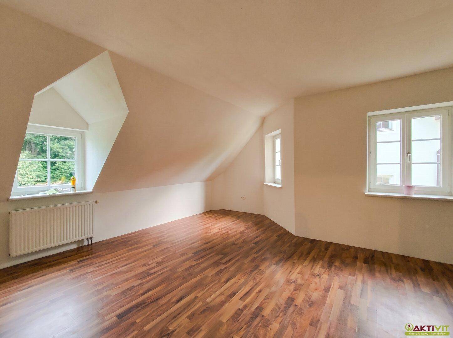 Heller Dachbodenraum mit schrägen Wänden, Holzboden und Ausblick ins Grüne.