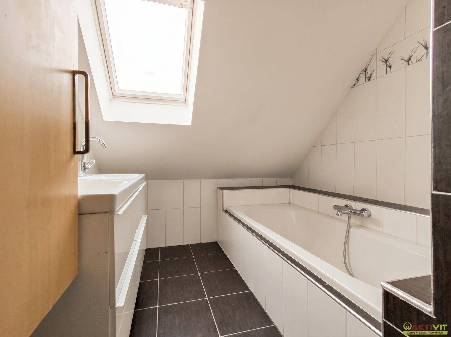 Badezimmer im Dachgeschoss mit Badewanne, Dachfenster und dunklen Bodenfliesen.