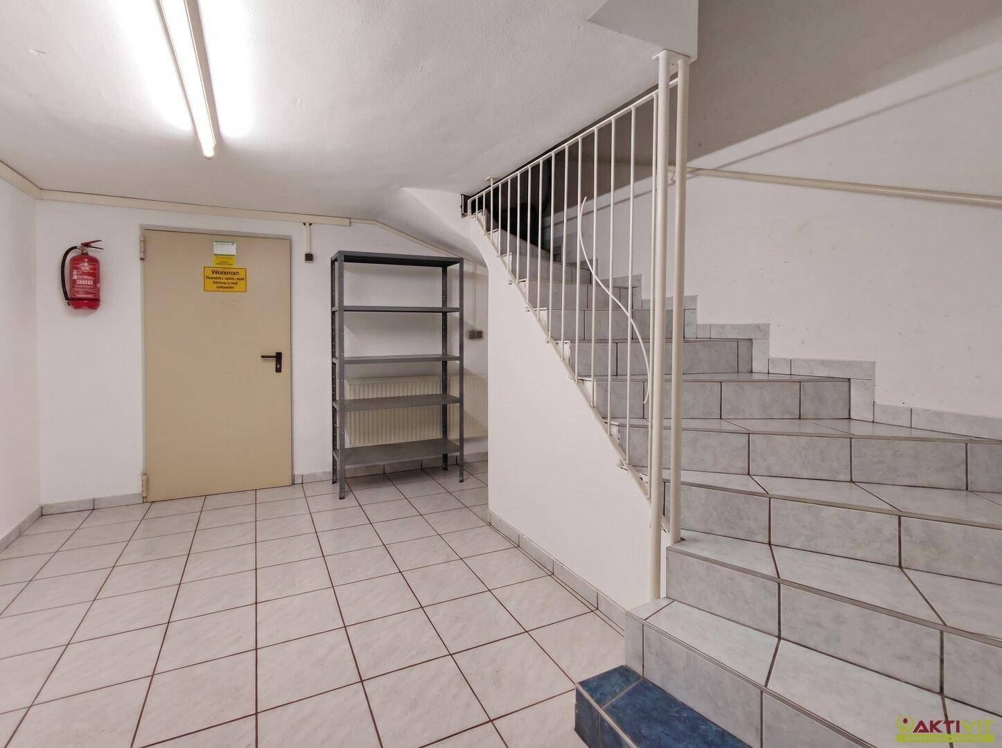 Kellerflur mit gefliestem Boden, Treppe und Brandschutztür, funktional gestaltet.
