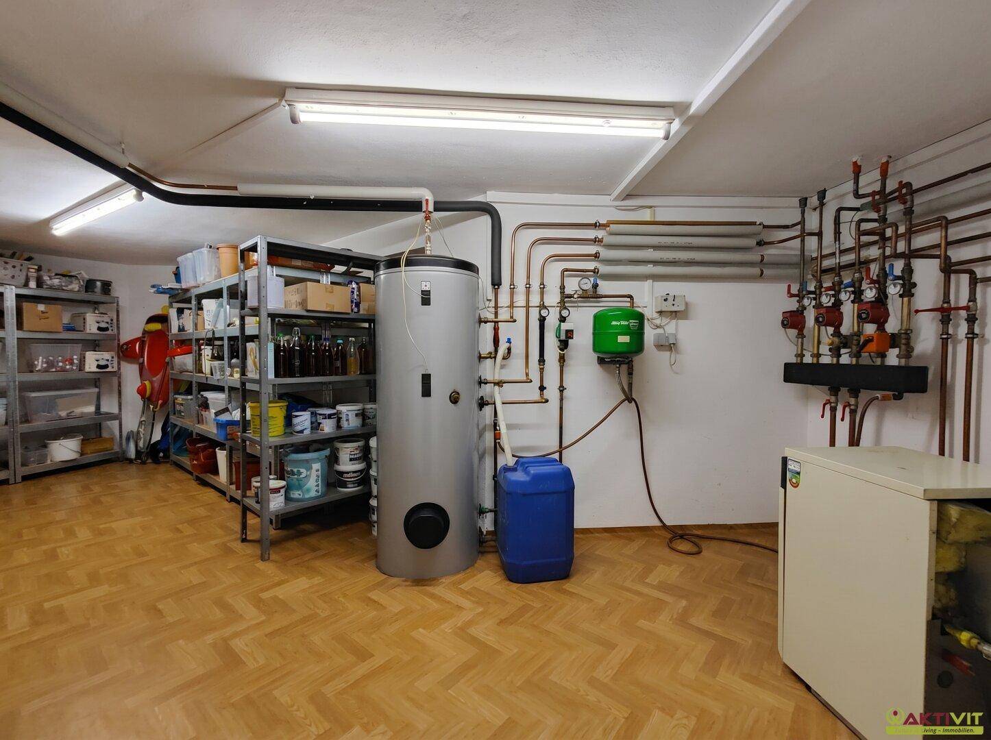 Heizungsraum im Keller mit Warmwasserspeicher, Rohrleitungen und Regalen für Lagerung.