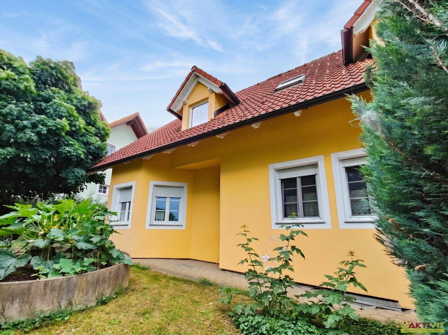 Gelbes Einfamilienhaus mit rotem Ziegeldach und gepflegtem Vorgarten unter blauem Himmel.