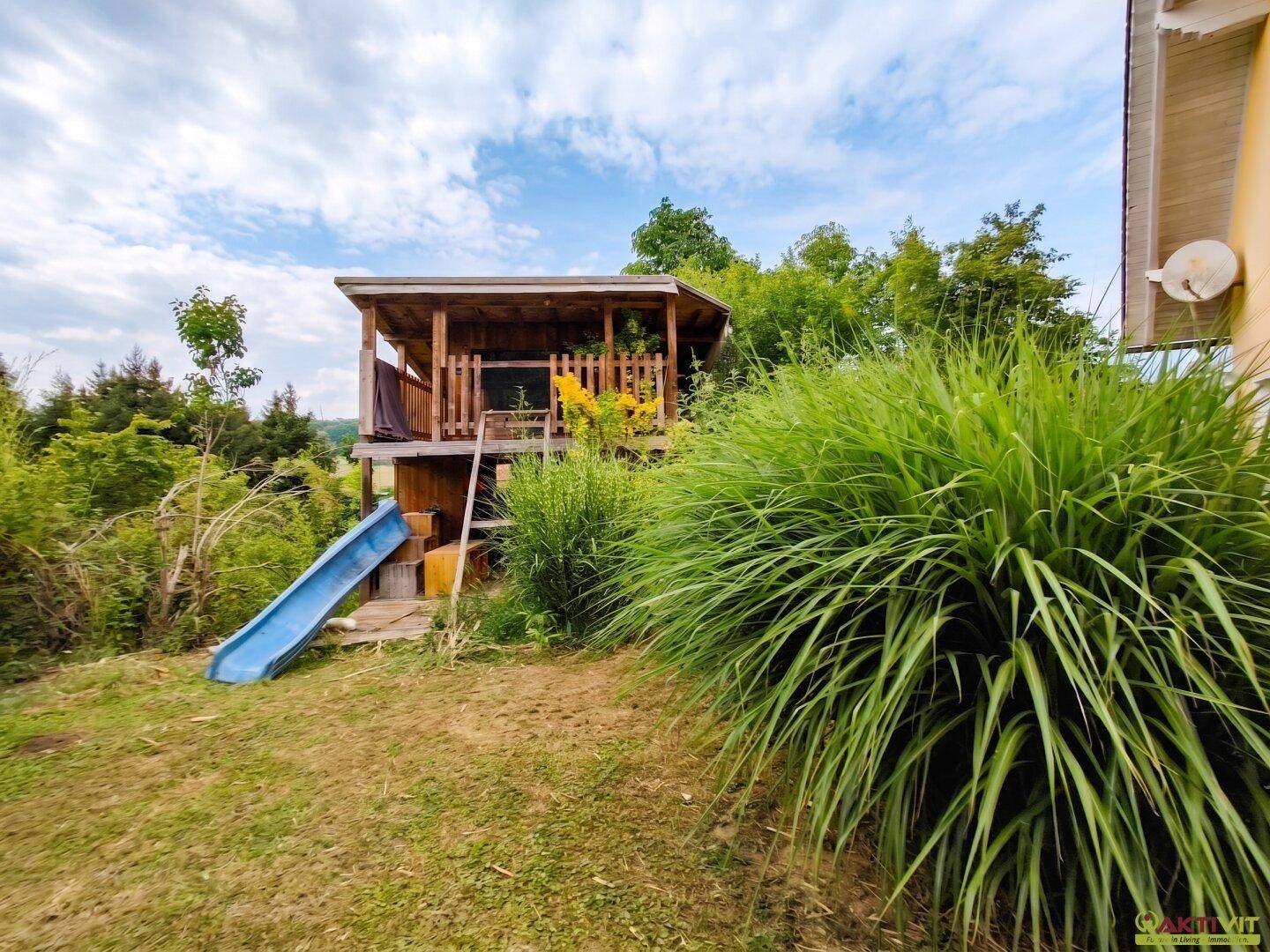 Großer Gartenbereich mit einem hölzernen Spielhaus und Rutsche, umgeben von üppigem Grün.