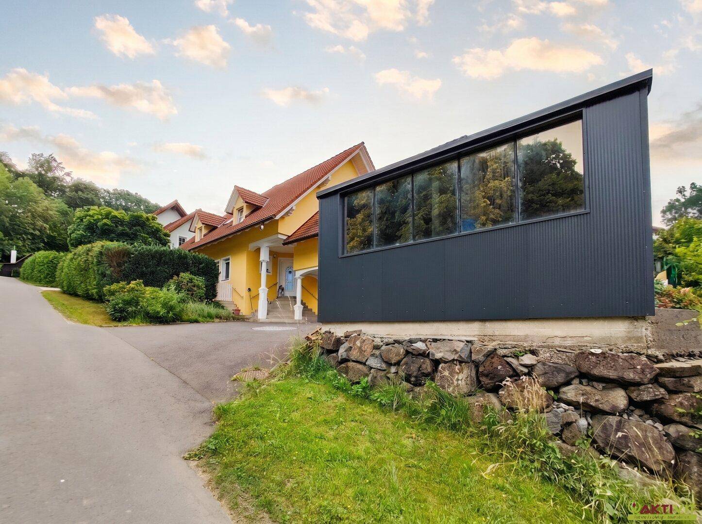 Einfamilienhaus mit gelber Fassade und modernem, dunklem Anbau, eingebettet in grüne Umgebung.