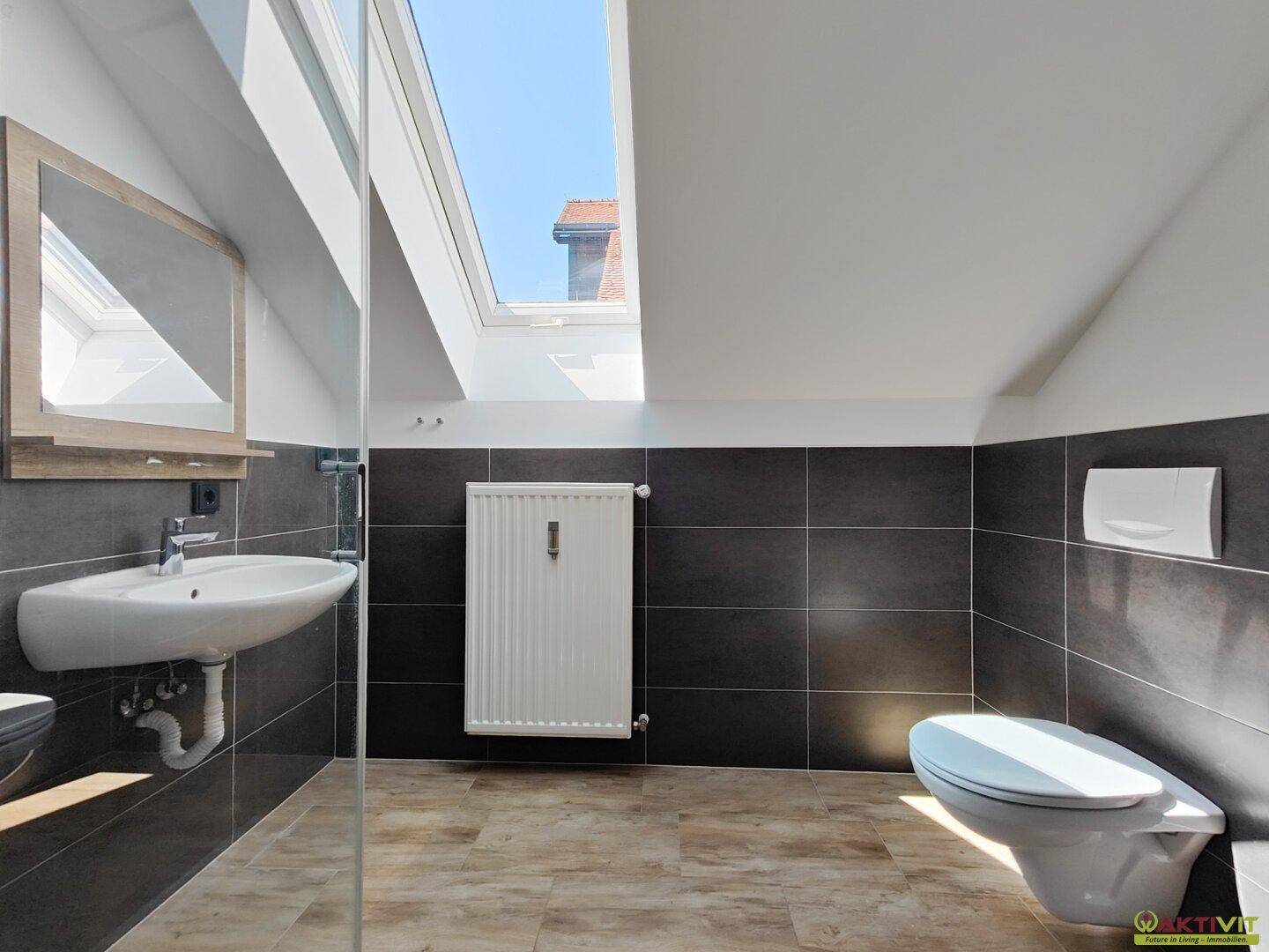 Modernes Badezimmer mit dunklen Fliesen, wandhängendem WC und großem Dachfenster.