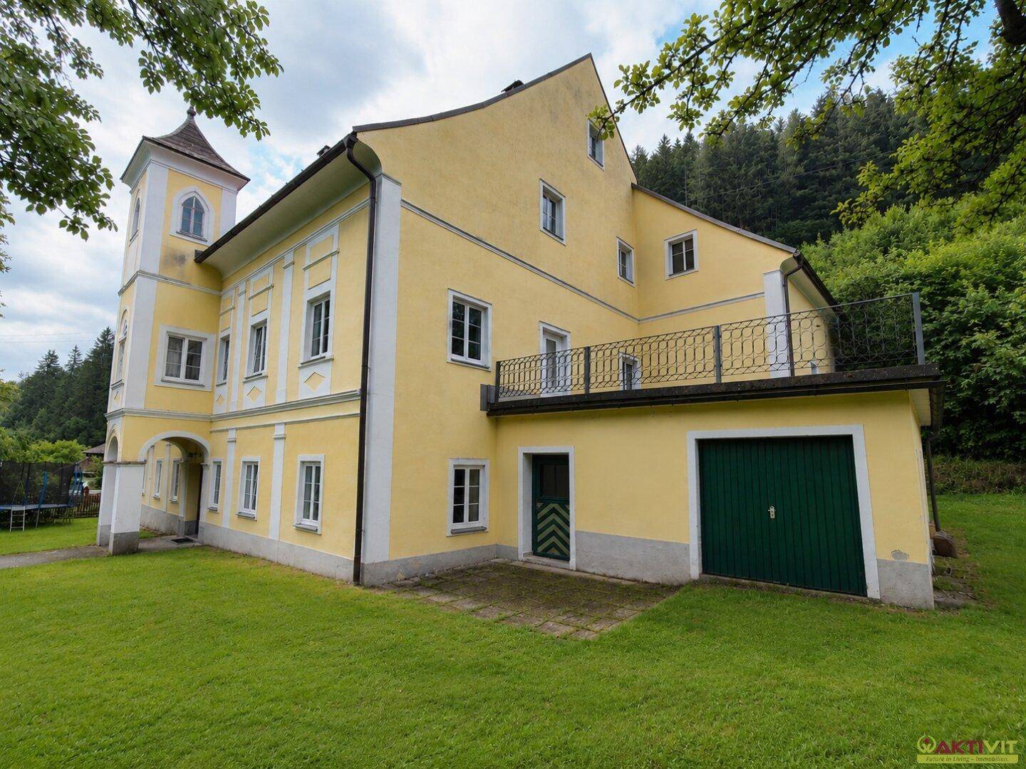 Seitenansicht des historischen Gebäudes mit Balkon, Garage und weitläufiger Grünfläche im Hintergrund.