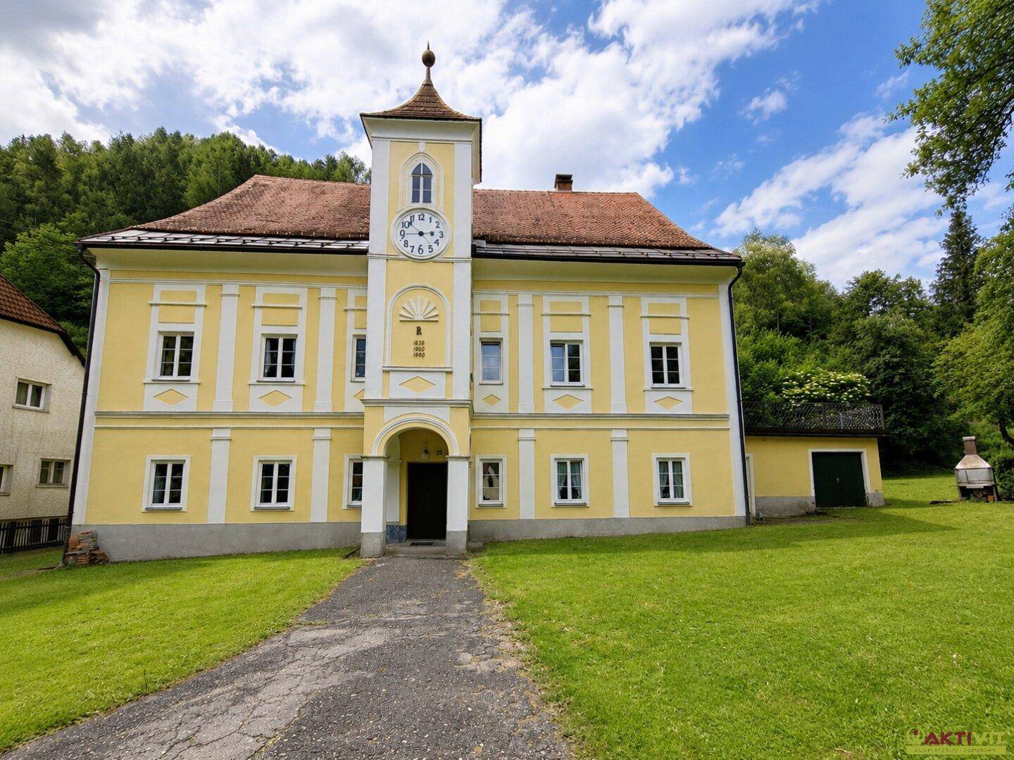 Frontansicht des historischen Gebäudes mit Uhrturm, rotem Dach und weitläufigem Rasenbereich.