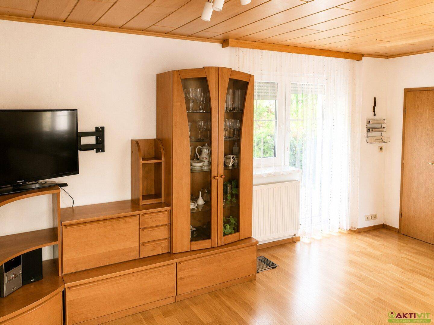 Wohnzimmer mit Holzmöbeln, Fernseher und Fenster mit hellem Vorhang.