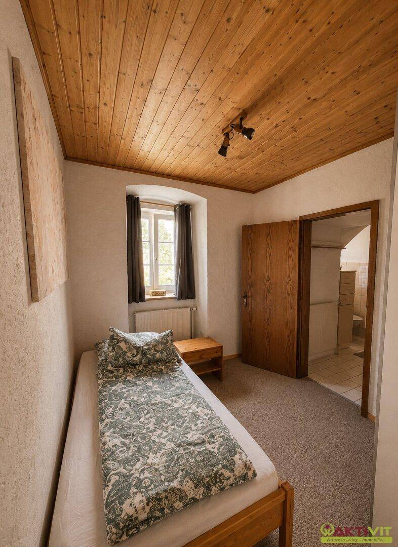 Schlafzimmer mit Einzelbett, Holzdecke und Zugang zu einem weiteren Raum.