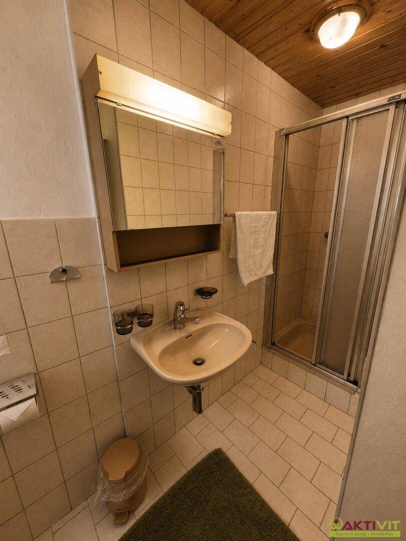 Badezimmer mit Dusche, Waschbecken und Spiegelschrank, gefliest in Beige.