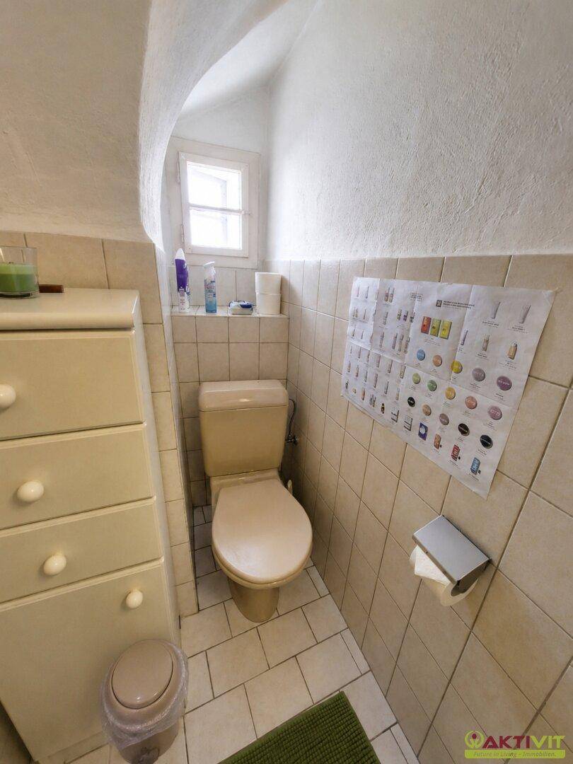 Separate Toilette mit Fenster, beigen Fliesen und kleinem Schrank.