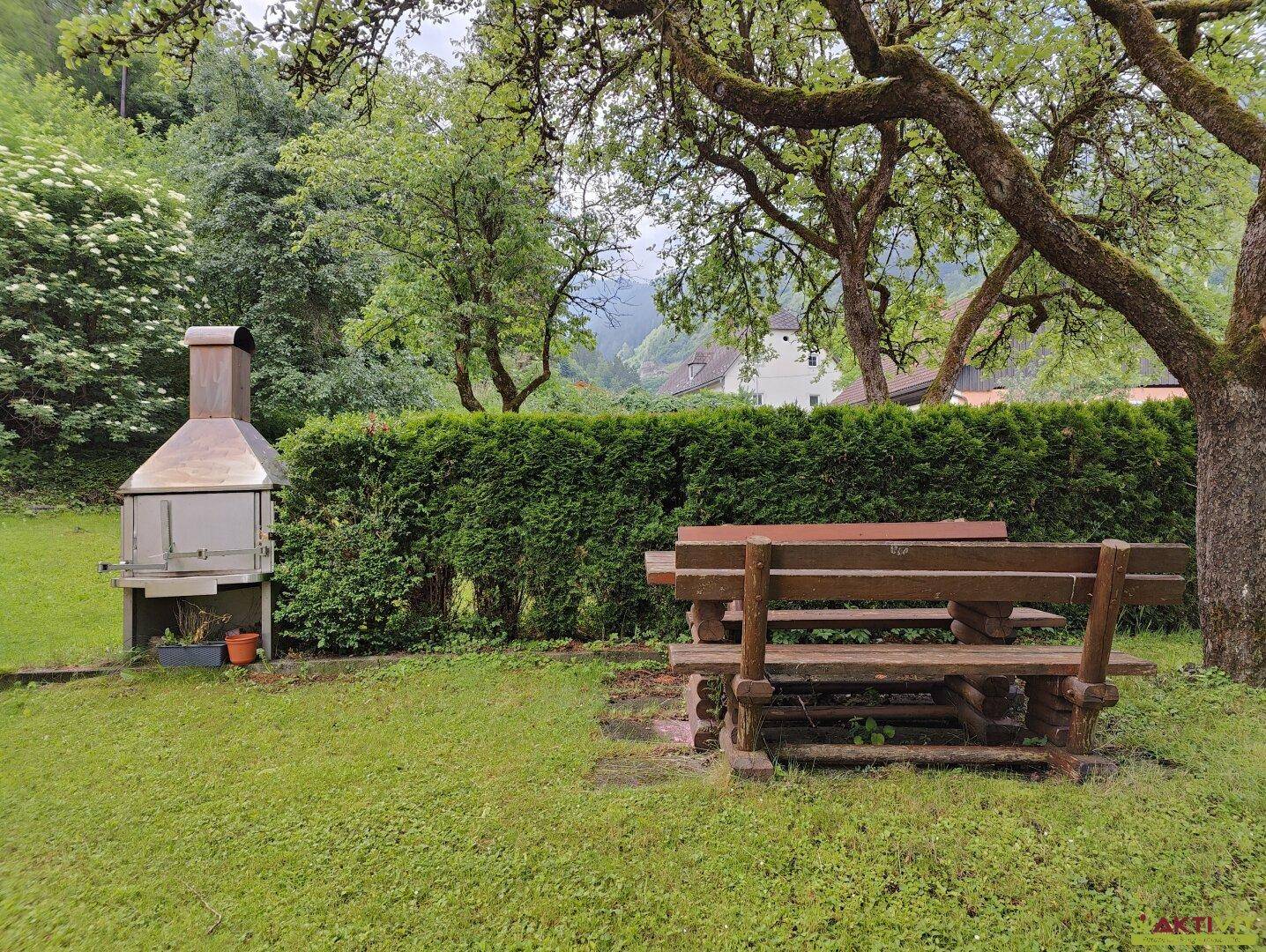 Gartenbereich mit Grillplatz, Holzbänken und üppiger Hecke im Hintergrund.