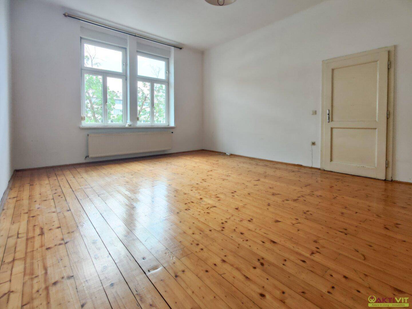 Großes, helles Zimmer mit glänzenden Holzdielen und zwei Fenstern, die viel Licht hereinlassen.