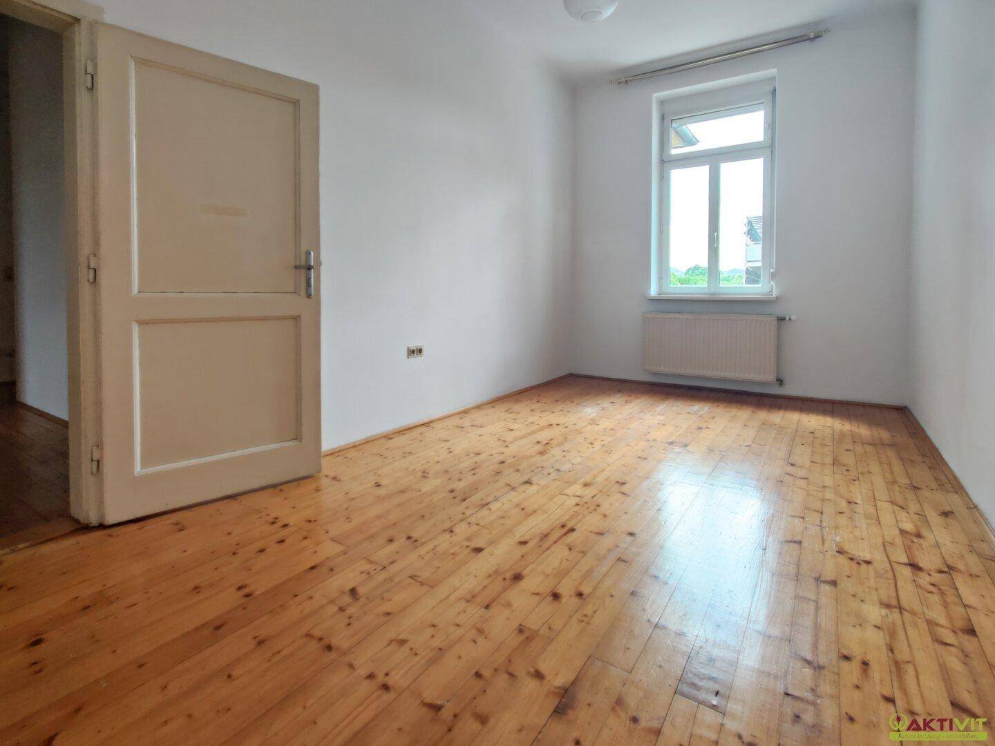 Helles Schlafzimmer mit Holzdielenboden und einem Fenster, das den Raum mit Tageslicht füllt.