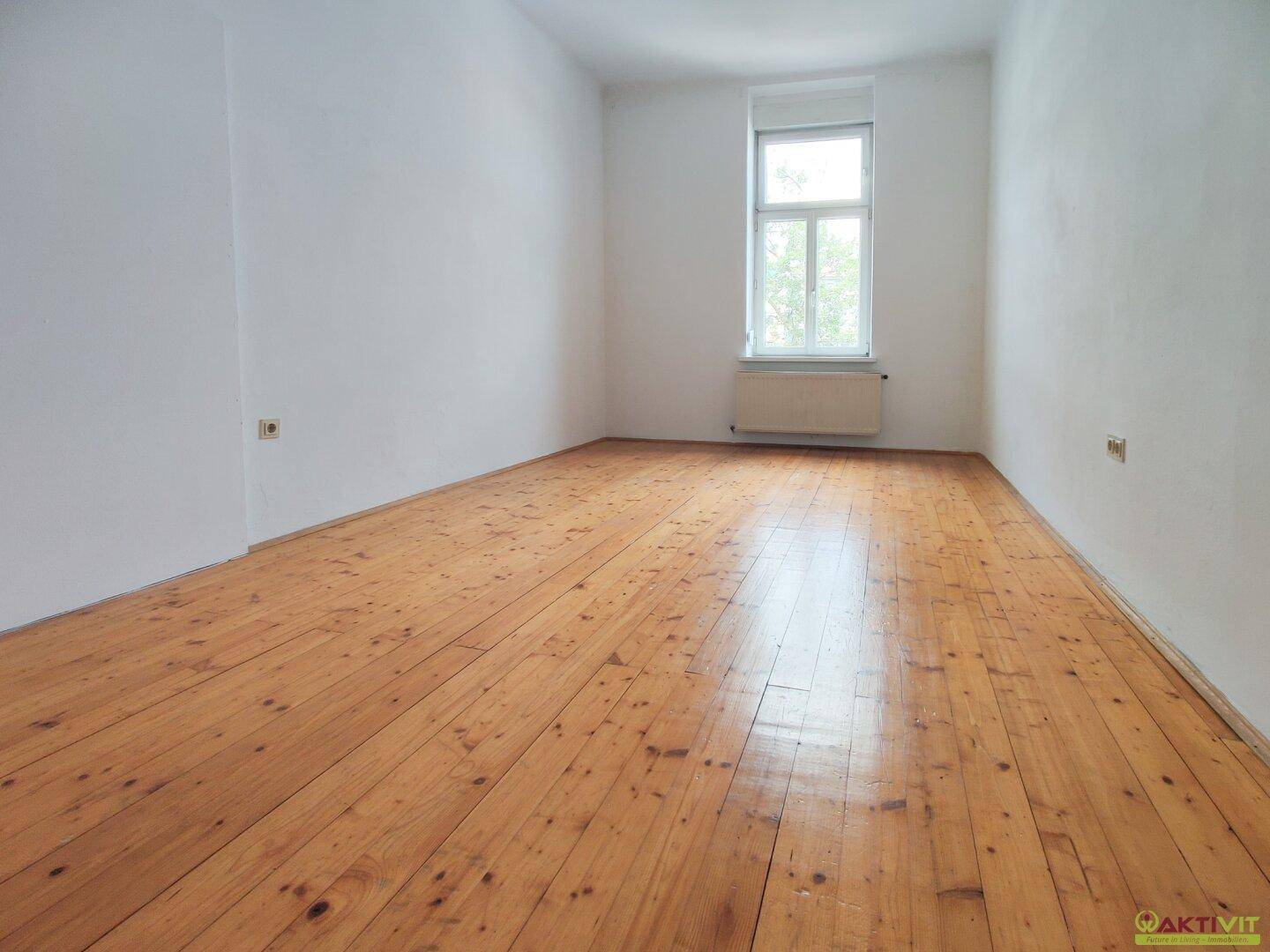 Leeres, helles Zimmer mit Holzdielenboden und einem Fenster, das für natürliche Belichtung sorgt.