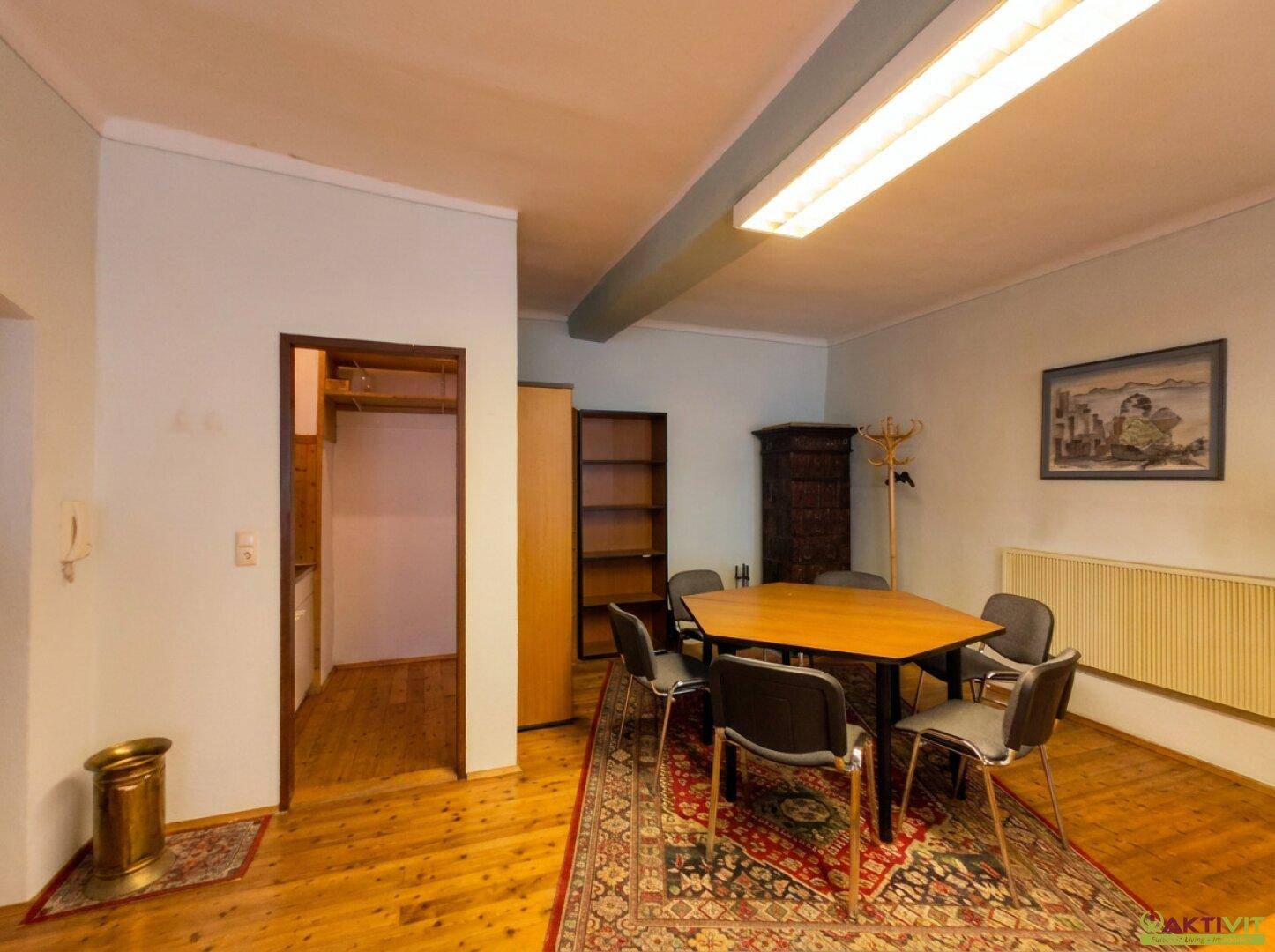 Besprechungsraum mit Holztisch, Stühlen und Teppich auf Holzboden, daneben ein offener Durchgang.