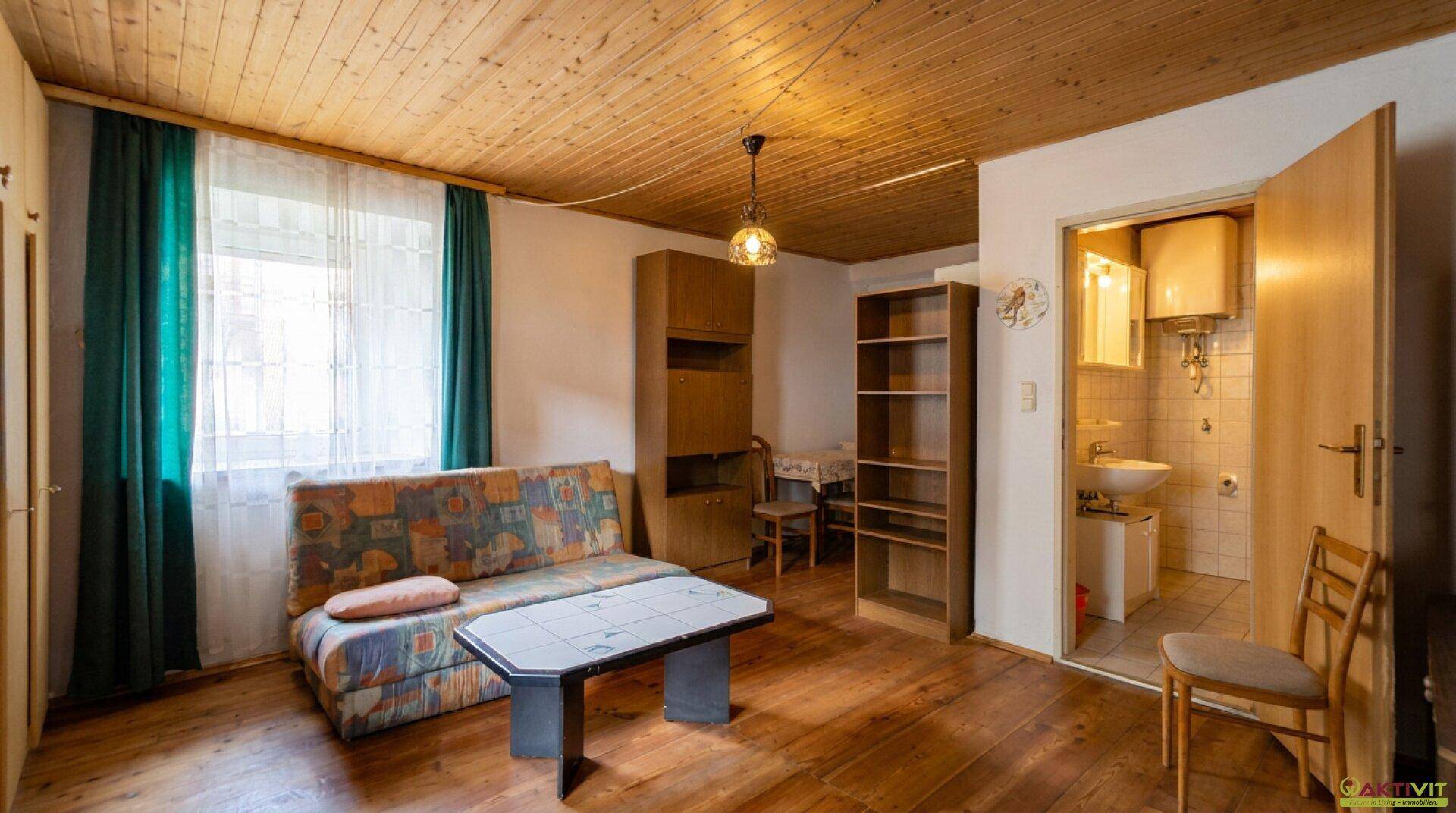 Gemütliches Wohnzimmer mit Schlafsofa, Holzboden und Holzdecke, daneben eine kleine Küchenzeile.