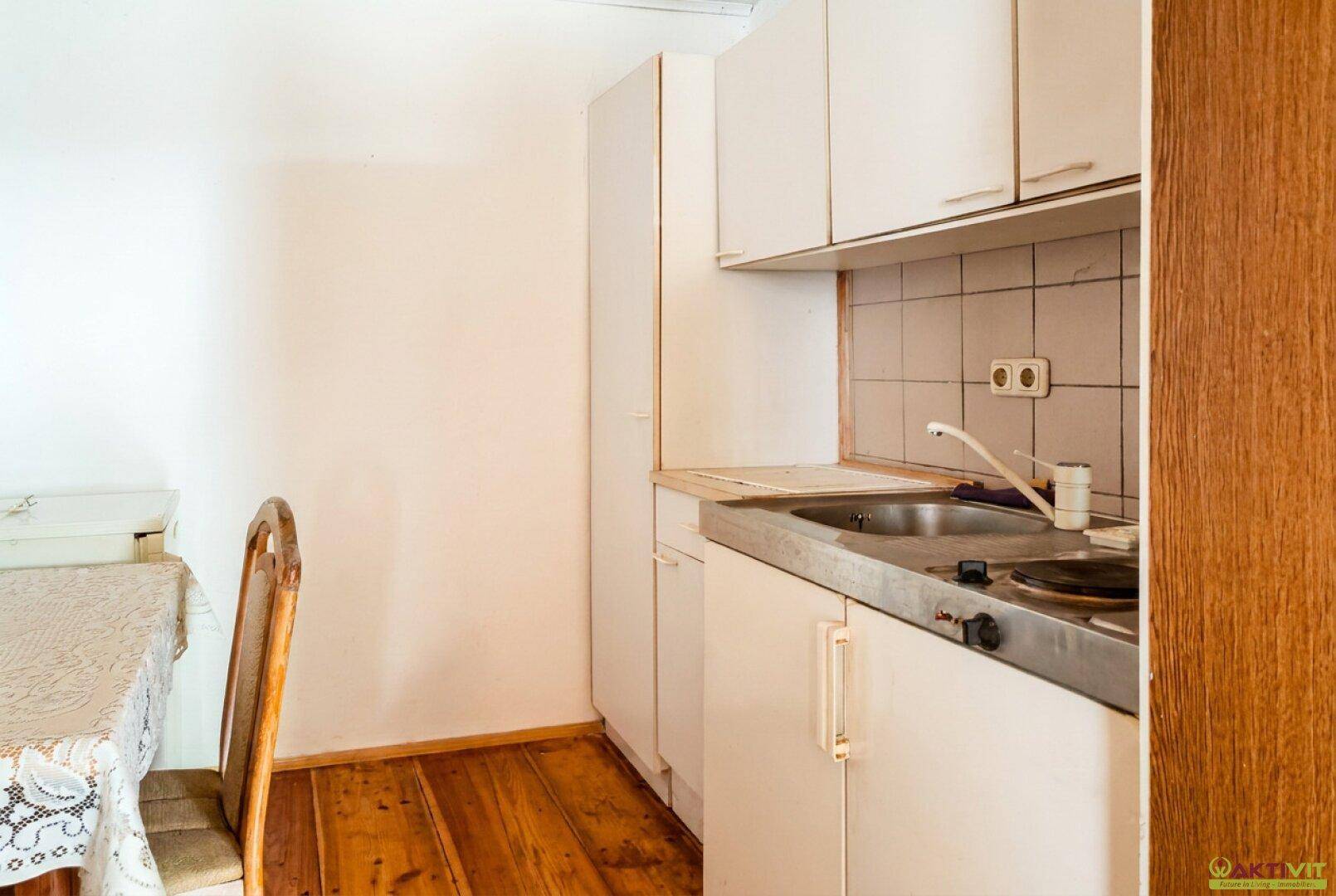 Einfache Küchenzeile mit Spüle, Kochfeld und Holzboden, daneben ein kleiner Esstisch.