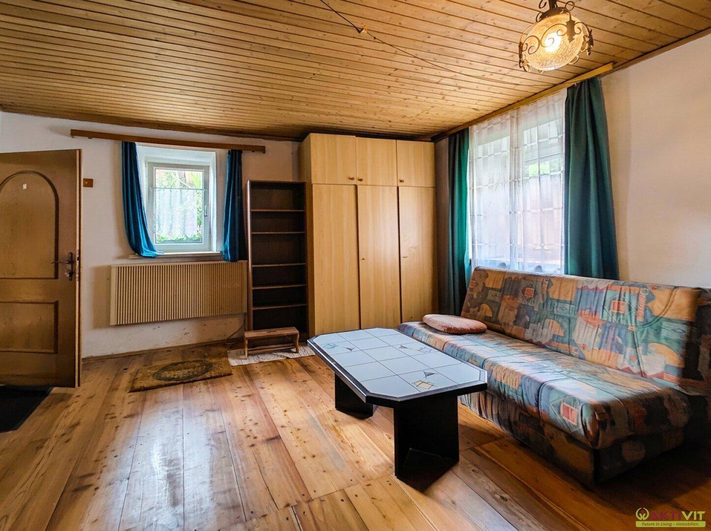 Wohnzimmer mit Schlafsofa, Holzboden, Holzdecke und großem Fenster mit Vorhängen.