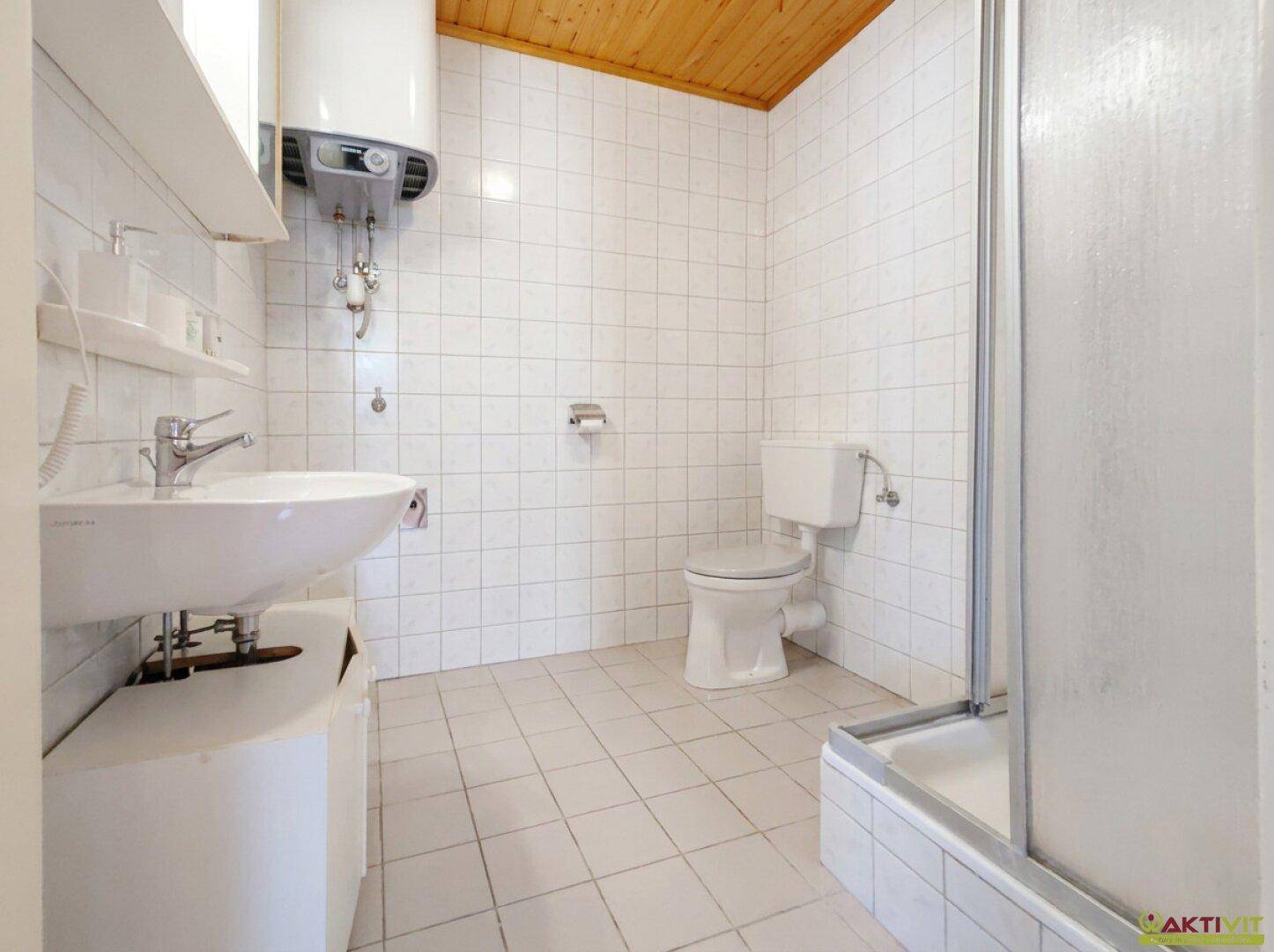 Badezimmer mit weißen Fliesen, WC, Waschbecken und Duschkabine, sowie Warmwasserboiler.