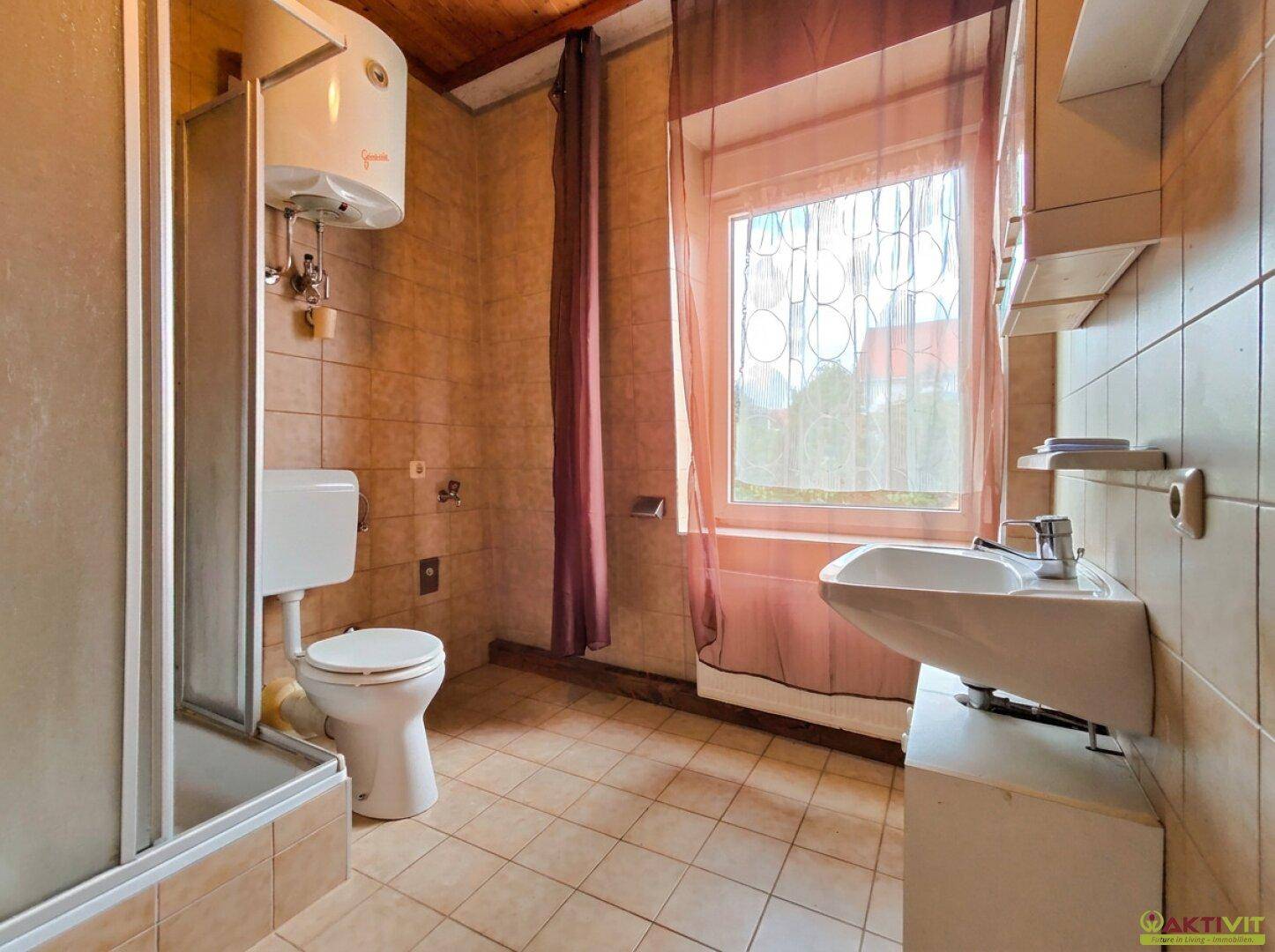 Badezimmer mit Fenster, Duschkabine, WC und Waschbecken, alles in beigen Fliesen gehalten.