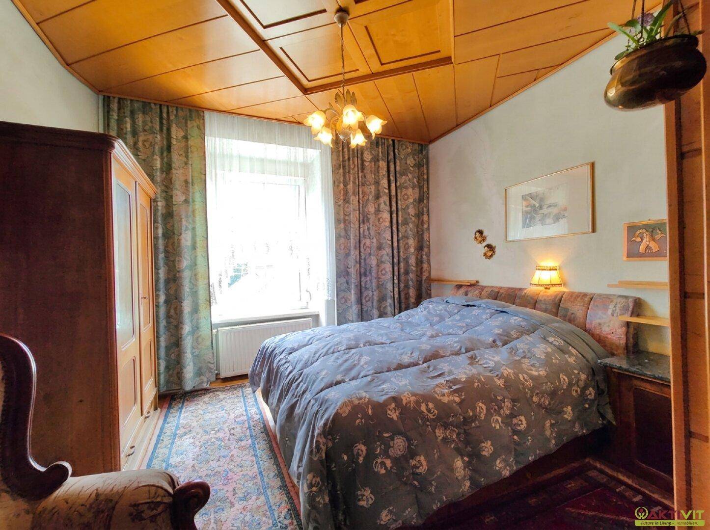 Schlafzimmer mit großem Bett, Holzdecke und traditioneller Einrichtung, hell durch Fenster.