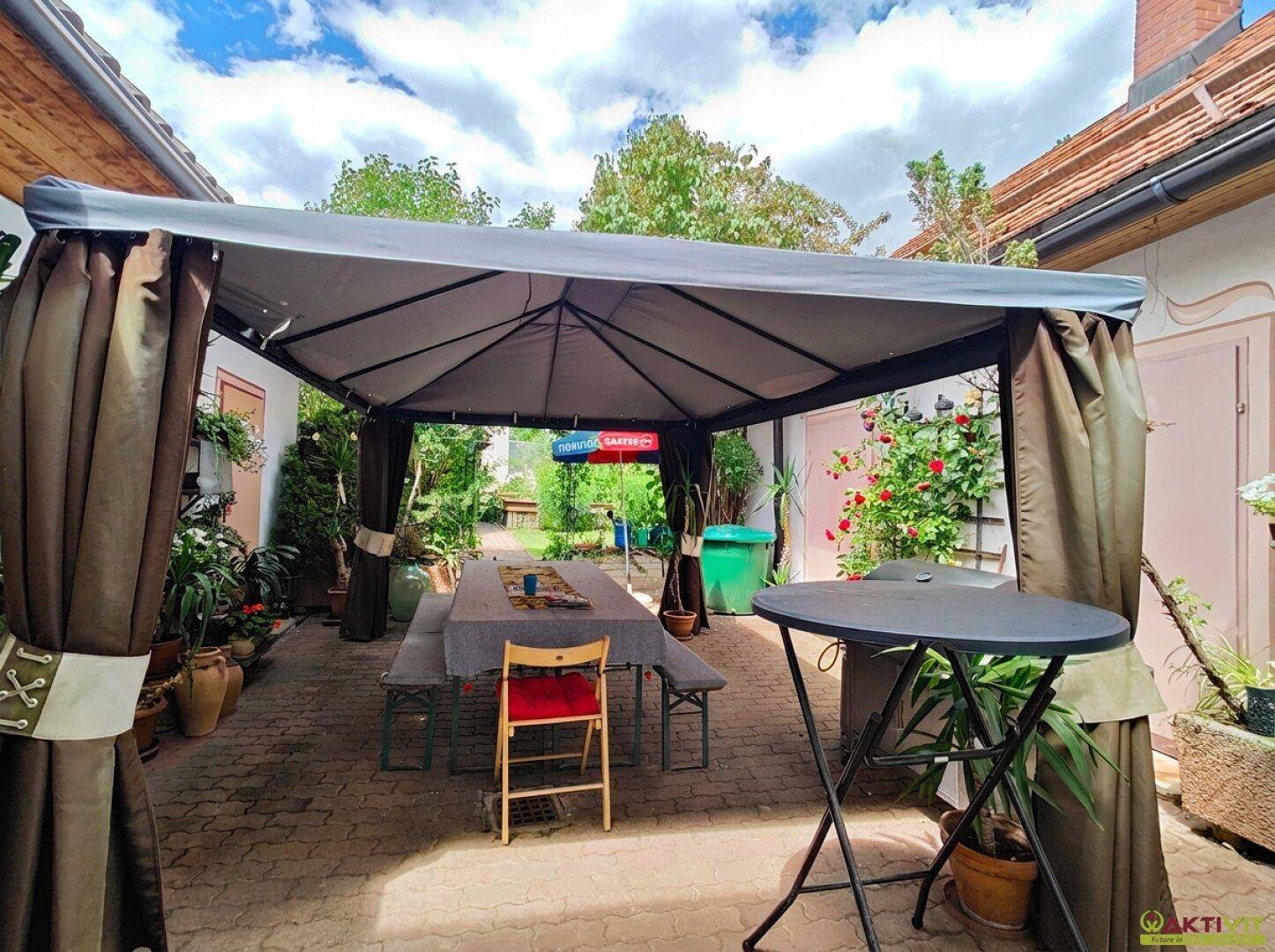 Überdachte Terrasse mit Esstisch und Sitzbänken, umgeben von Pflanzen im Freien.