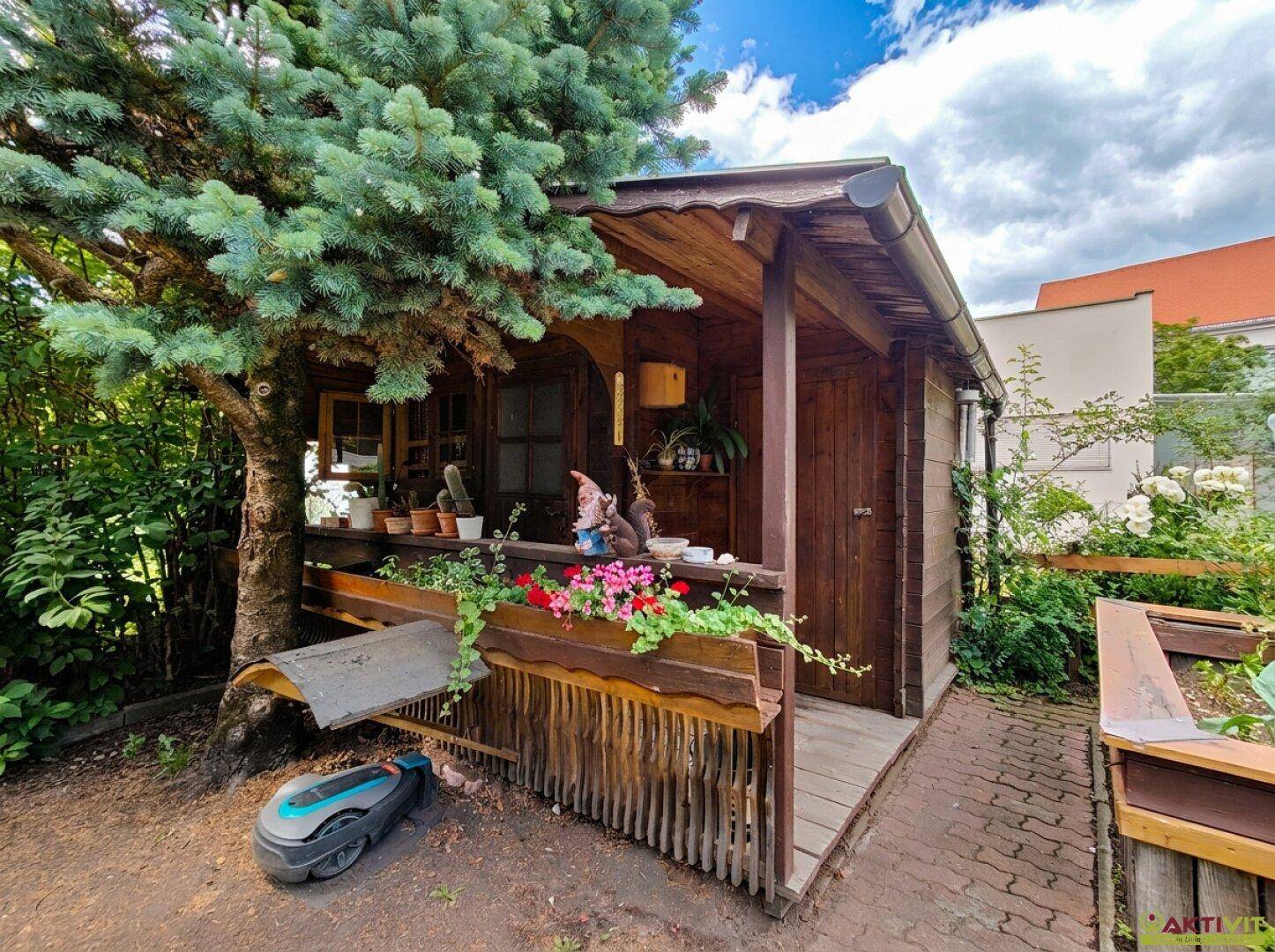 Rustikales Gartenhaus aus Holz mit kleiner Veranda und Blumenkästen unter einem Baum.