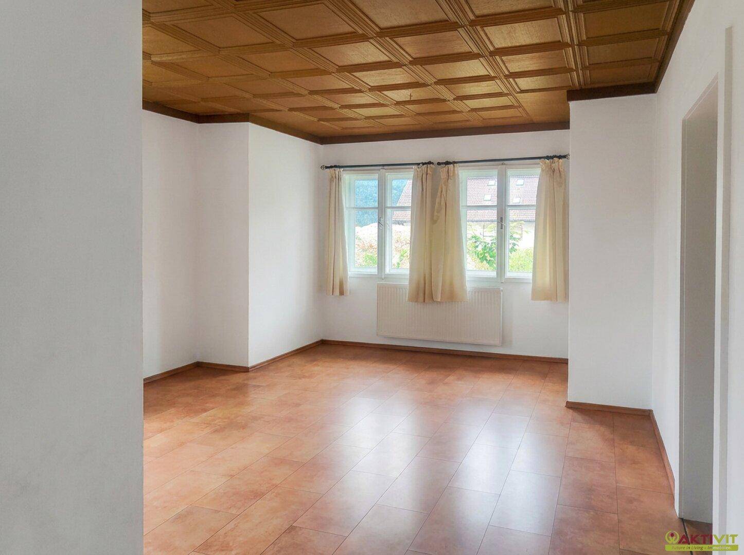 Leeres Wohnzimmer mit Holzdeckenverkleidung, Fliesenboden und Blick ins Grüne.