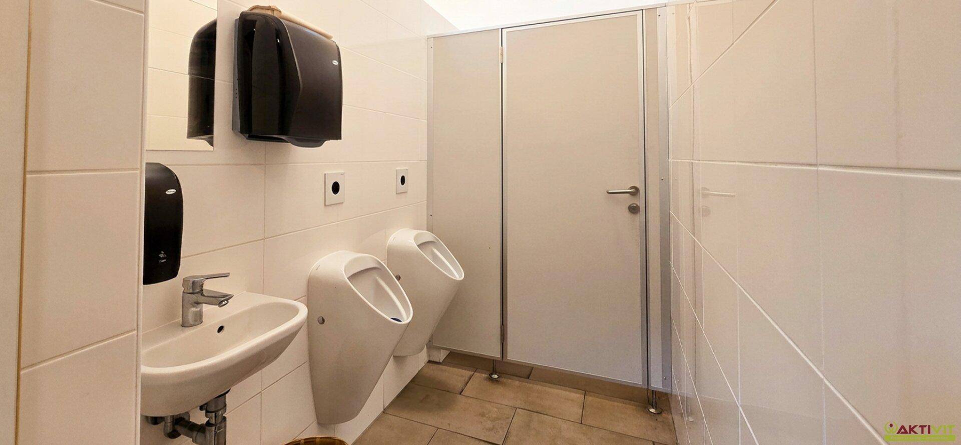 Öffentlicher Toilettenraum mit Waschbecken und zwei Urinalen, komplett mit weißen Wandfliesen.