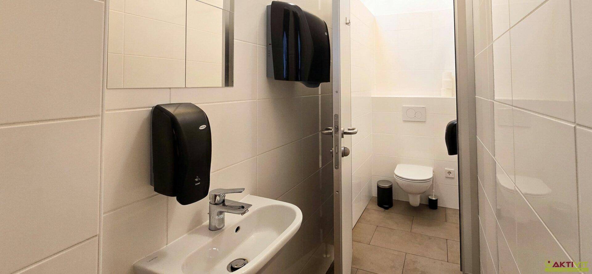 Sauberer öffentlicher Toilettenraum mit Waschbecken, Spiegel und WC, ausgestattet mit weißen Fliesen.