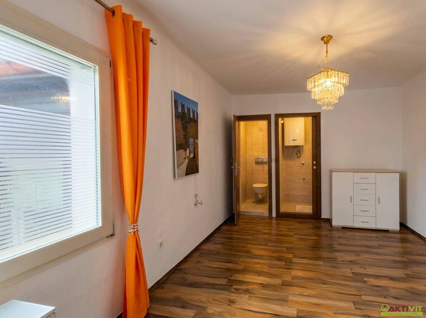 Heller Raum mit Holzboden, Kommode und Zugang zu Bad und WC.